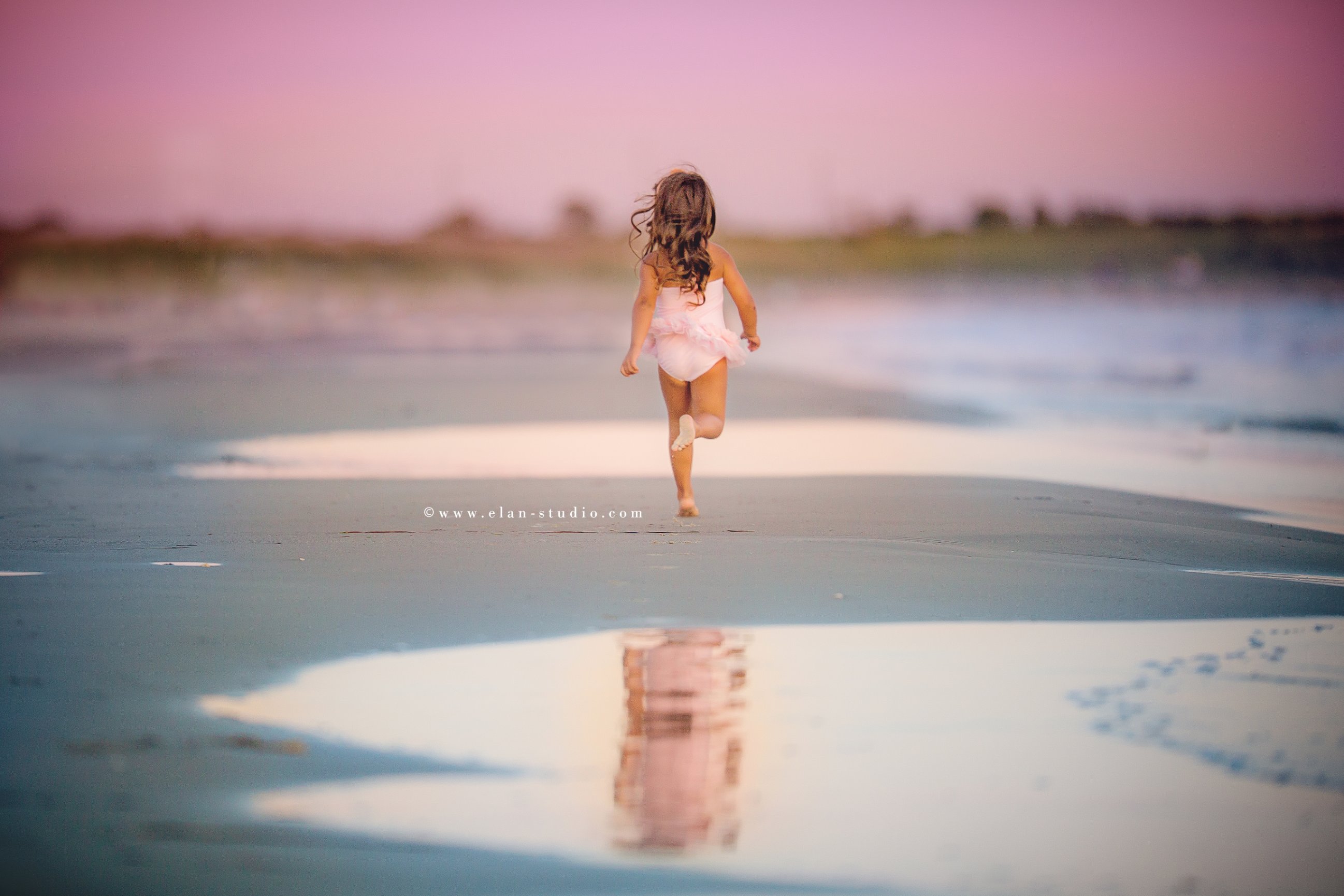little girl with curly brown hair running away on beach, with her image reflected in pool of water