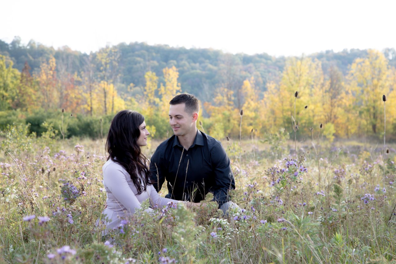 A couple sitting in a field of wildflowers with trees and hills in the background.