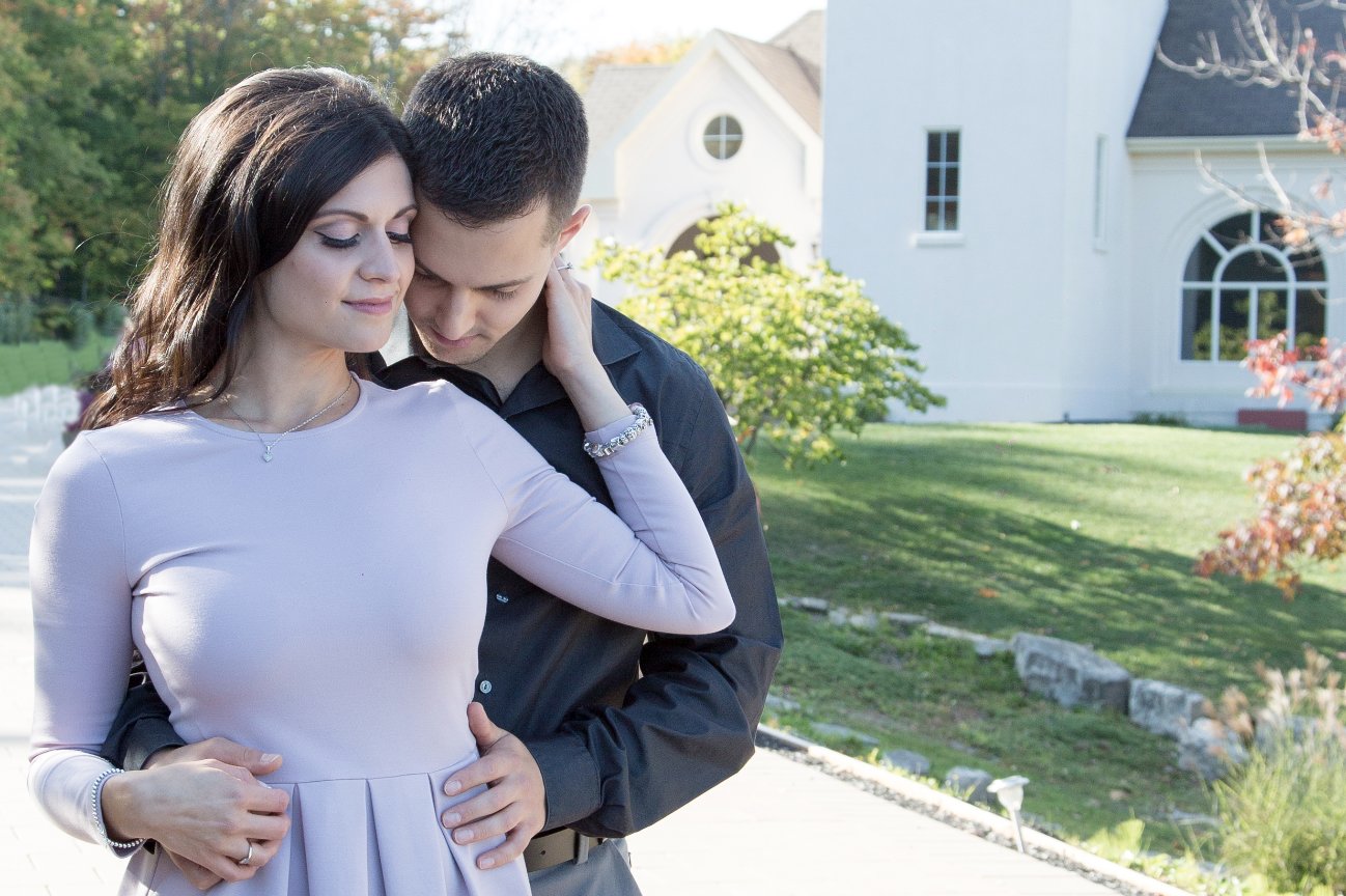 A couple embraces outdoors near a white building, surrounded by greenery and trees.