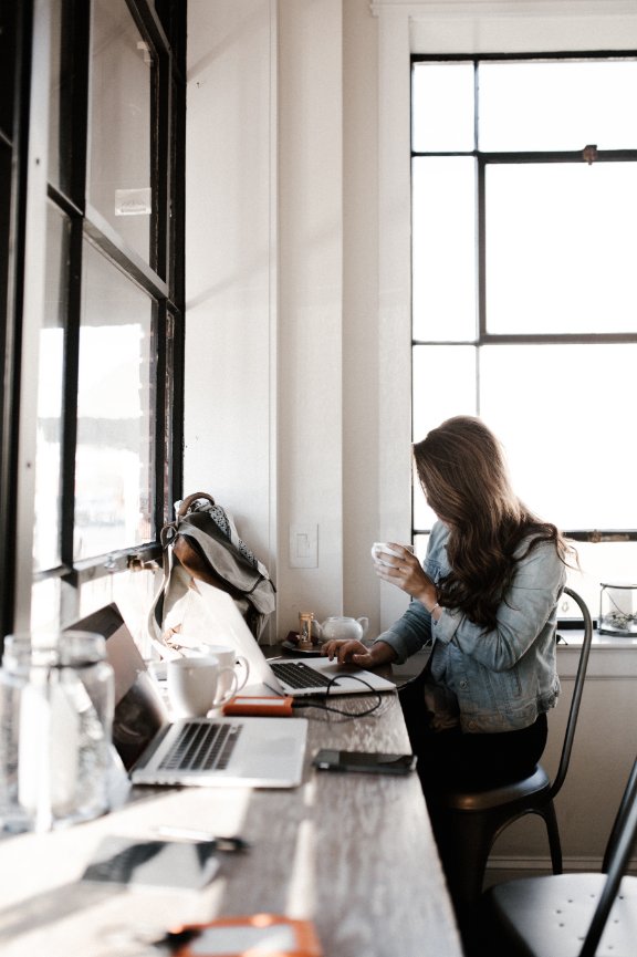 A person working at a rustic desk with a laptop and coffee near a large window.