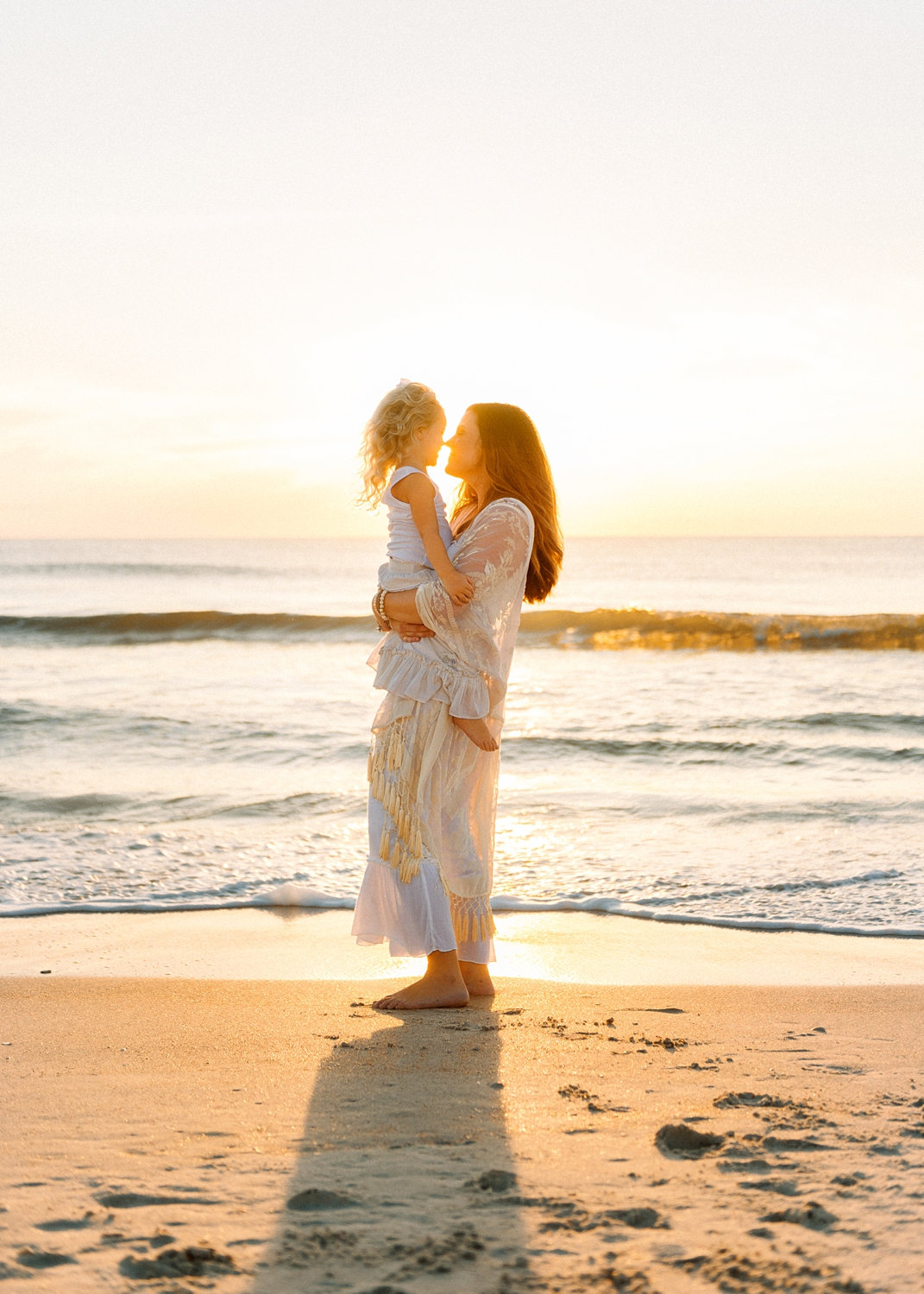 mom holding her young daughter, they are touching noses and silhouetted by the ocean sunrise behind them