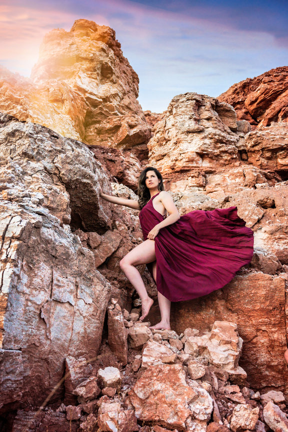 Woman in a flowing maroon dress poses on rocky terrain under a colorful sky.