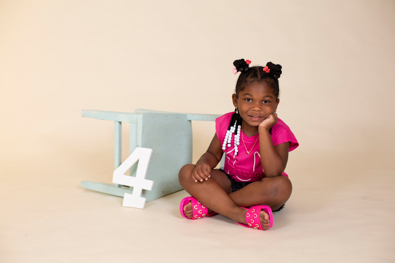 Baby girl sitting on a beige studio floor beside a number four prop.