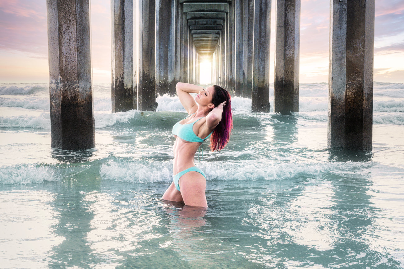 Woman in swimwear poses in shallow water under a pier at sunset, with waves gently splashing around.
