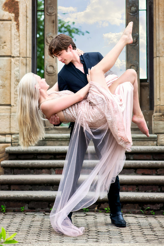 A man elegantly lifts a woman in a flowing pink dress, set against an ornate stone staircase and sky backdrop.