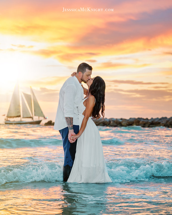 Couple sharing a romantic kiss in the ocean at sunset, with a sailboat in the background.