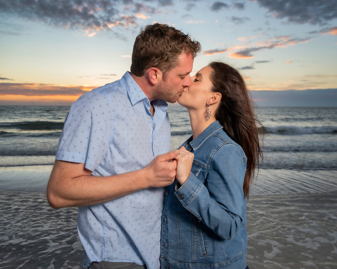 A couple shares a kiss on the beach at sunset, with gentle waves in the background.