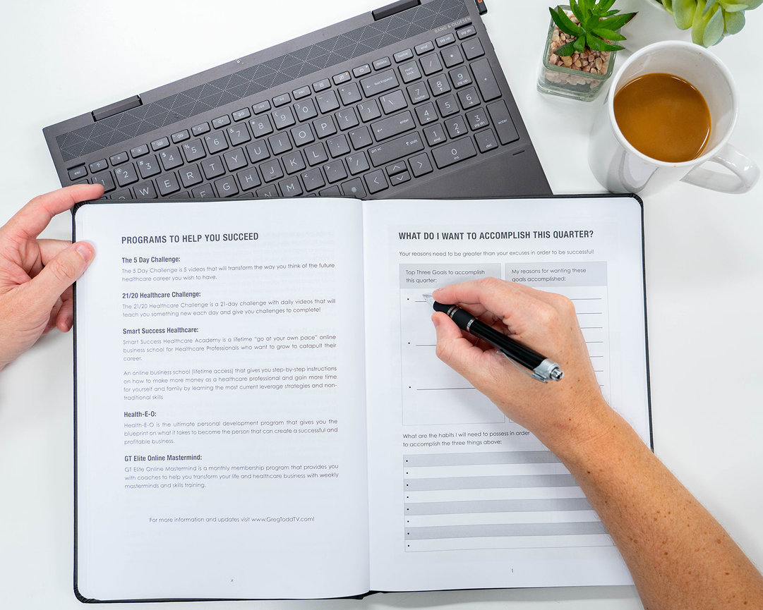A person's hand writing in a planner, with a laptop, coffee mug, and a succulent plant nearby.