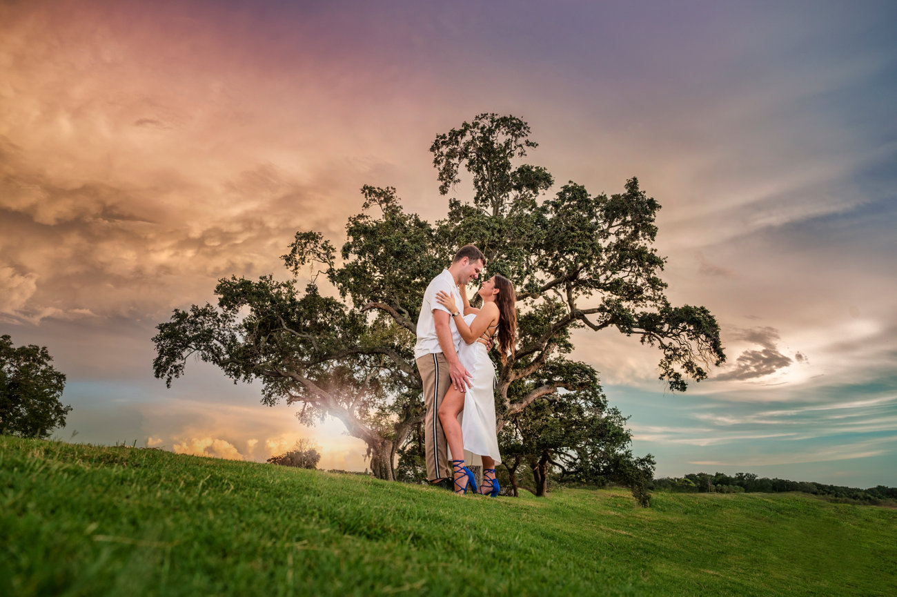 Couple embracing in a lush green field at sunset, with dramatic skies and a large tree in the background.