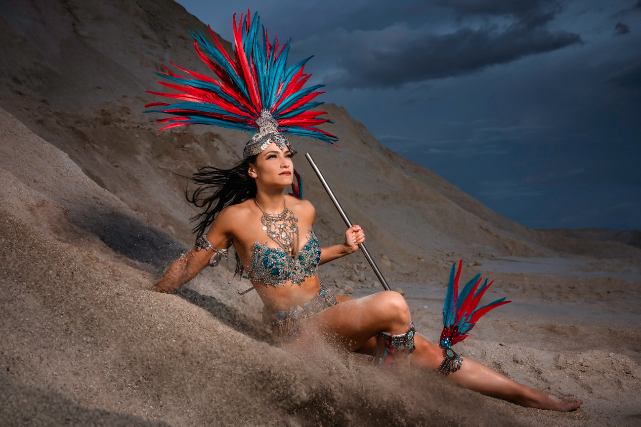 Dancer in vibrant costume with a feather headdress, seated on sandy terrain against a dramatic sky.