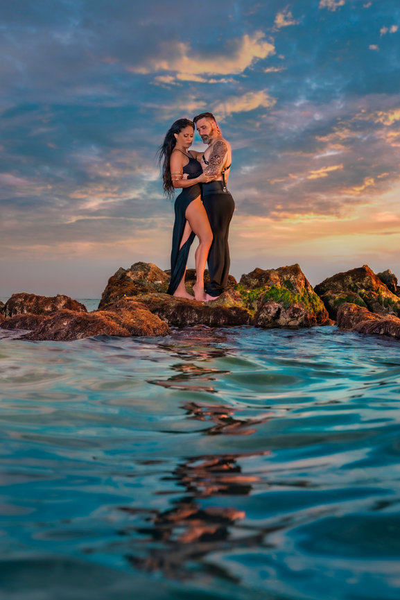 Couple embracing on rocky shore at sunset, with vibrant sky and calm water reflections.
