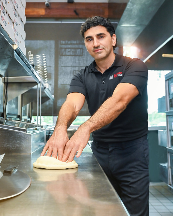 Man in a black shirt kneading dough at a pizzeria kitchen countertop.