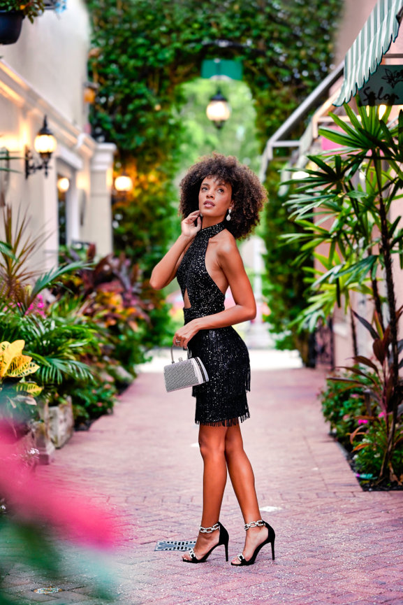 Woman in a black sparkly dress and heels poses in a lush garden pathway with vibrant plants.