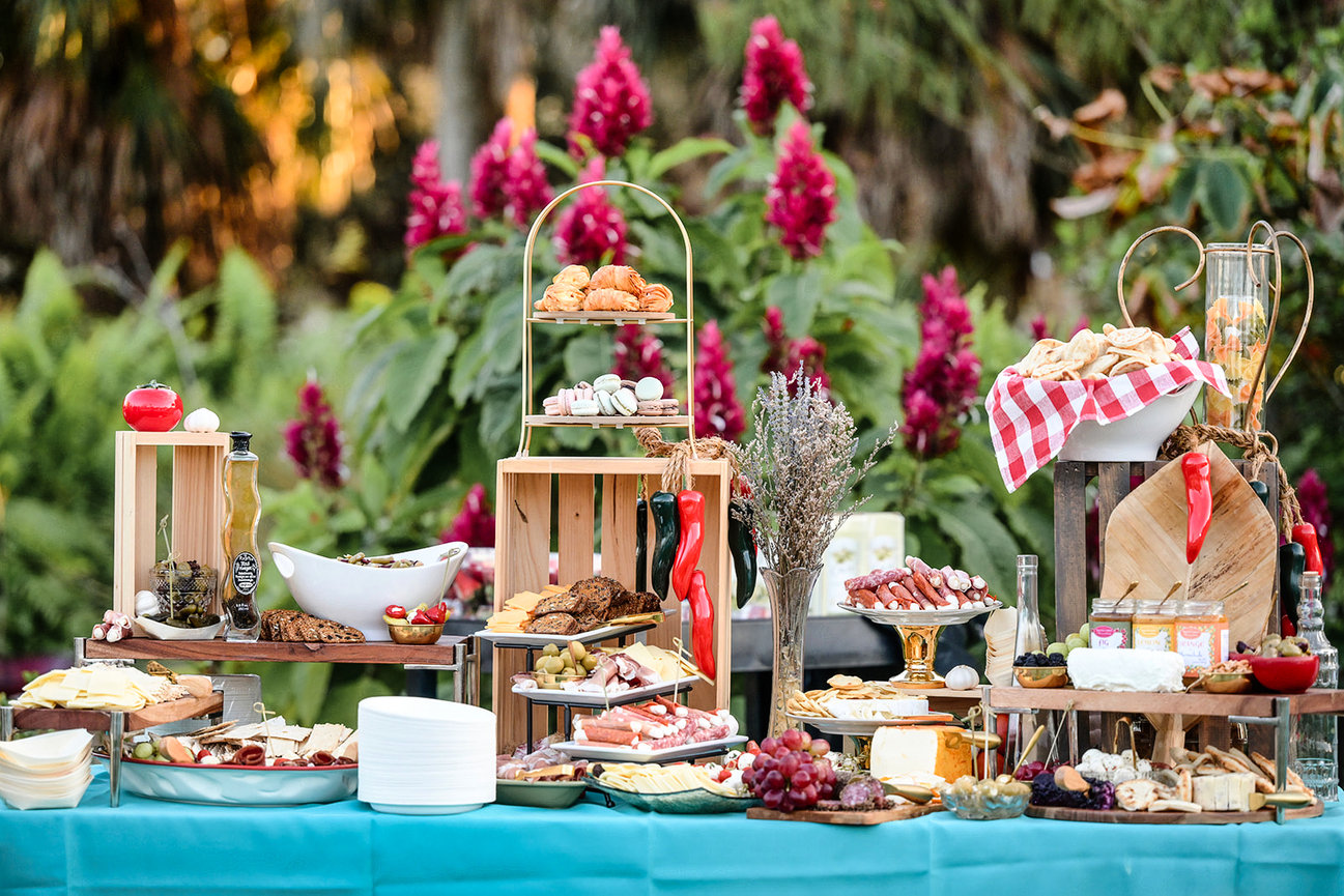 Elegant outdoor buffet featuring a variety of cheeses, pastries, fruits, and decorative flower arrangements.