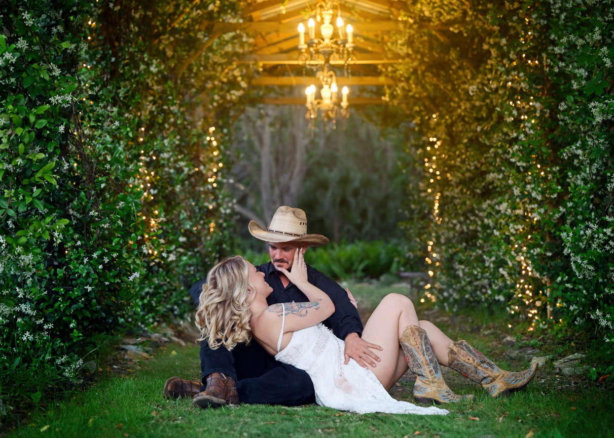 Couple with cowboy hat and boots in romantic outdoor setting, surrounded by greenery and soft lights.