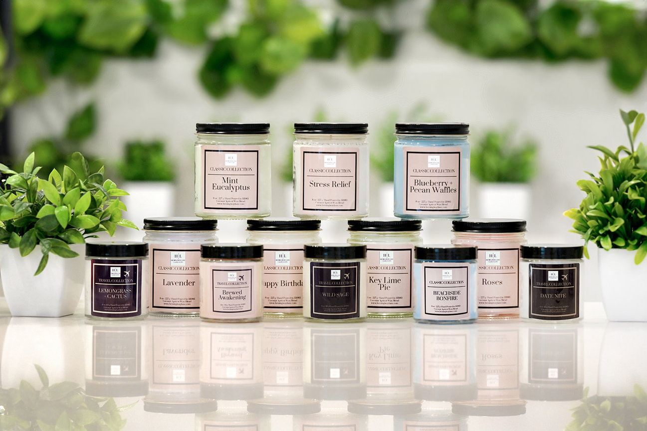 A display of scented candle jars arranged on a table, surrounded by small potted plants.
