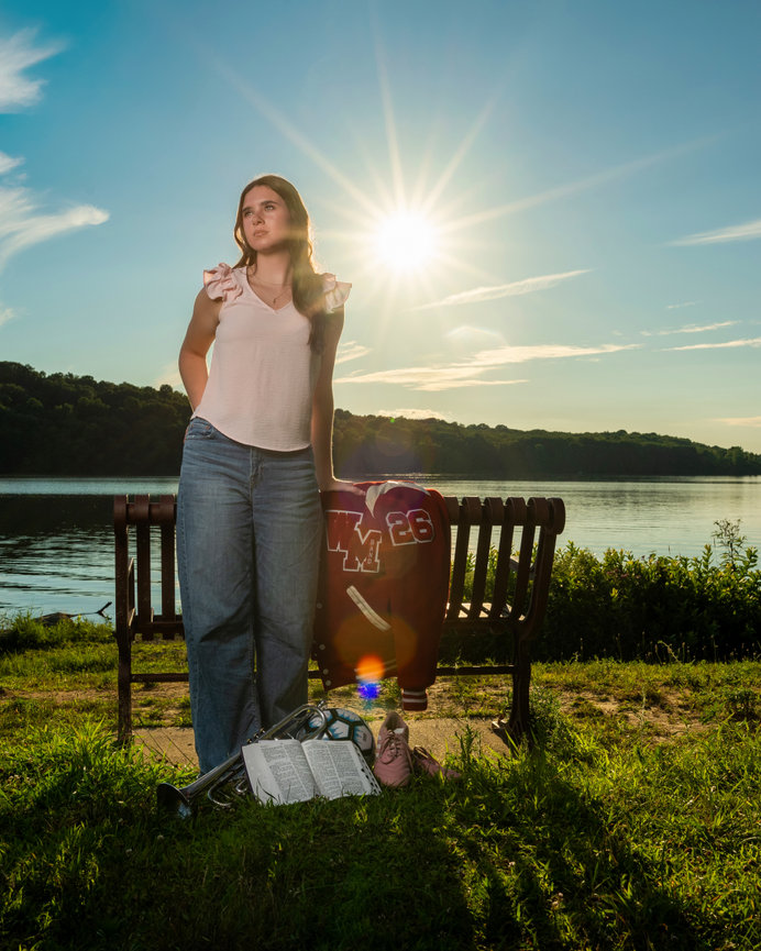 senior girl with her band letterman jacket, bible, trumpet and soccer shoes in front of a lake