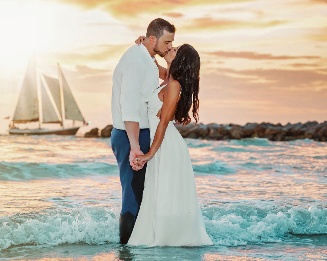 Couple kissing in the ocean at sunset, wearing wedding attire, with a sailboat in the background.