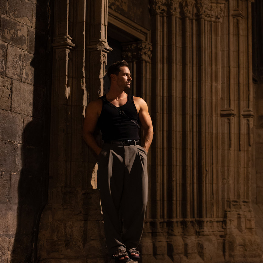 Man in dark tank top and trousers stands against ornate stone wall at night, hands in pockets, looking to the side. Barcelona night. Professional ballroom dancer. Paso Doble