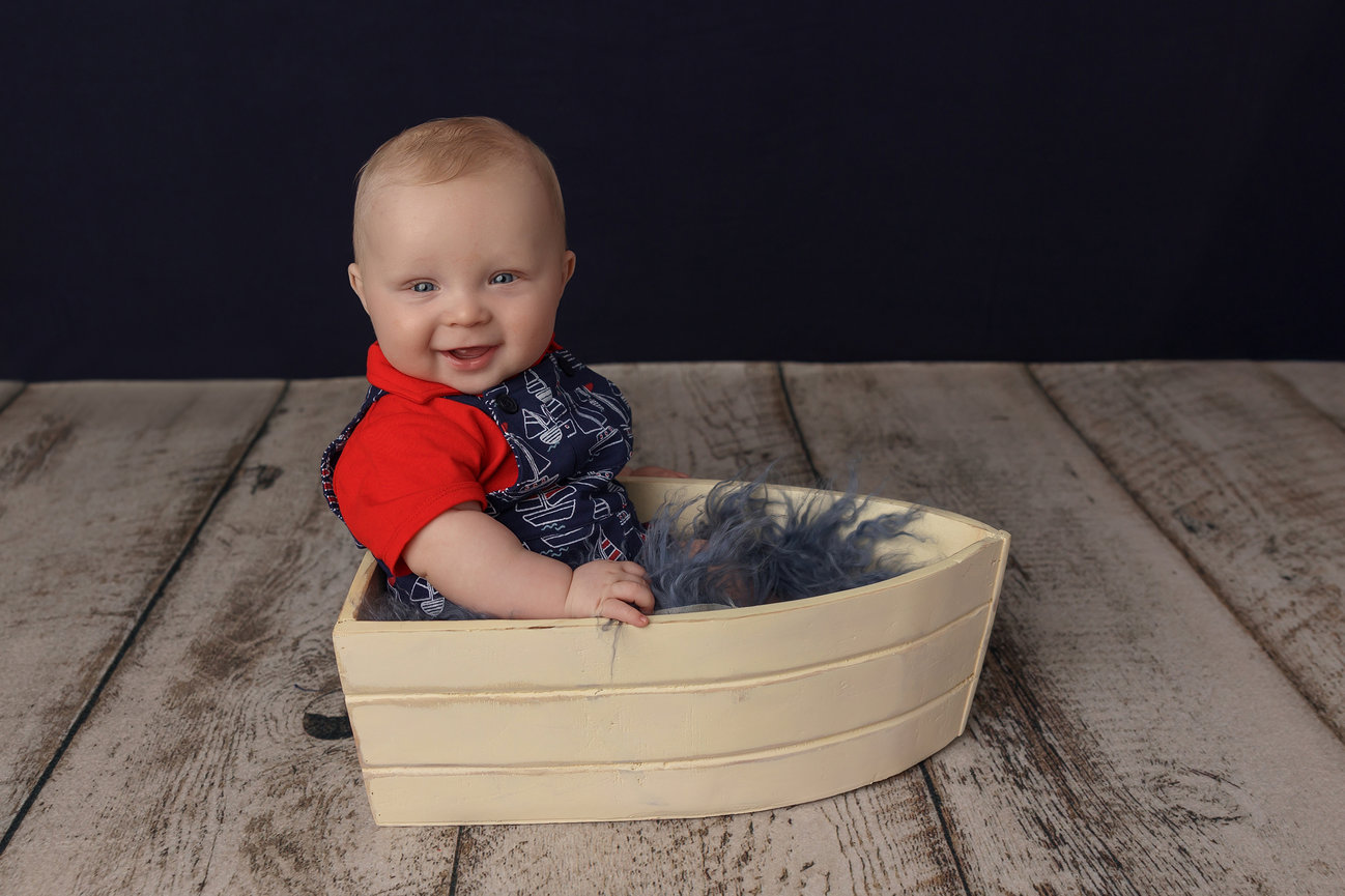 A smiling baby in a toy boat on a wooden floor, wearing a red shirt and navy overalls.
