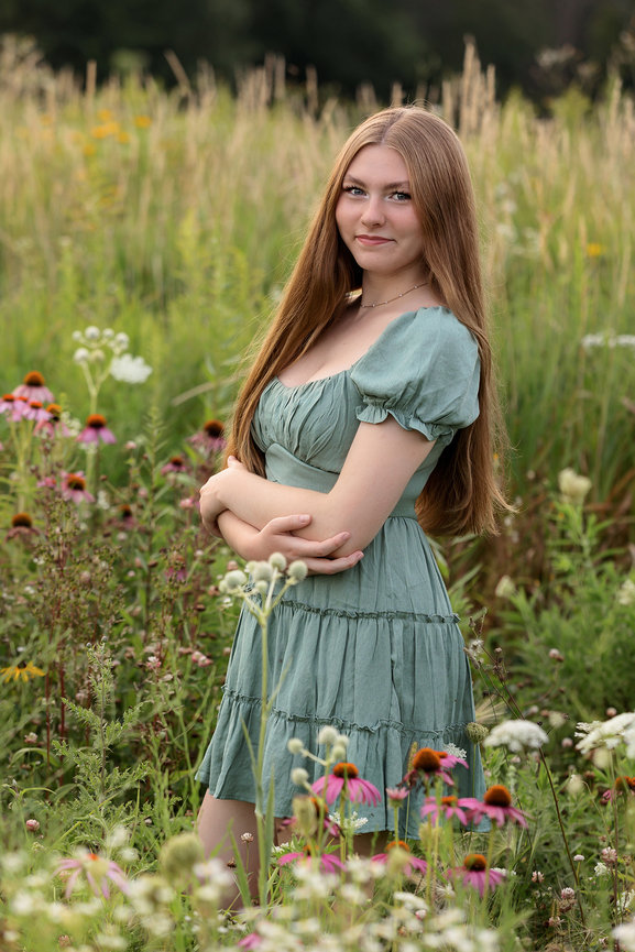 Young woman in a green dress standing in a wildflower field, with arms crossed and long hair flowing.