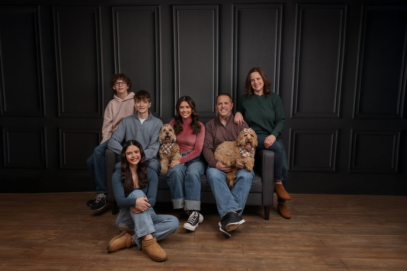 Family of six with two dogs sitting on a couch, posed against a dark paneled wall.