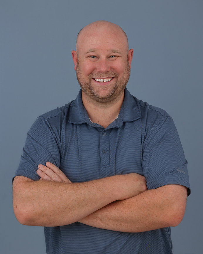 Bald man smiling with arms crossed, wearing a blue polo shirt against a gray background.