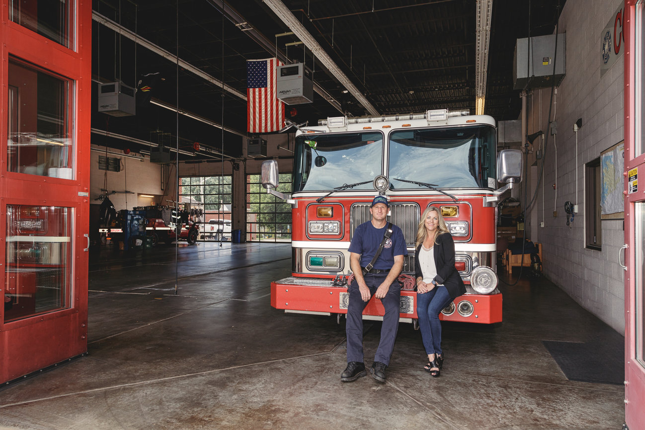 Firefighter and woman sitting on a fire truck in a station with an American flag overhead.