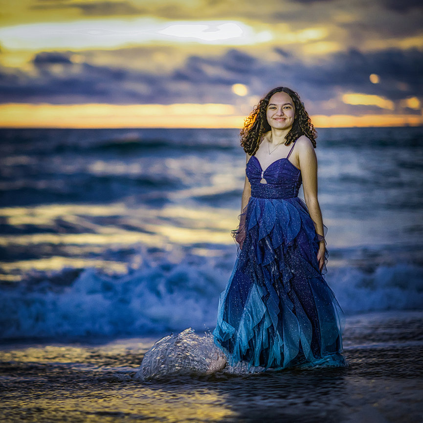 Galveston Beach senior portrait at sunset, girl in long flowing blue fairytale dress at the ocean's edge with dramatic Texas Gulf sunset reflections, ethereal cinematic master-level photography by Ty Swartz Swartz Portraits
