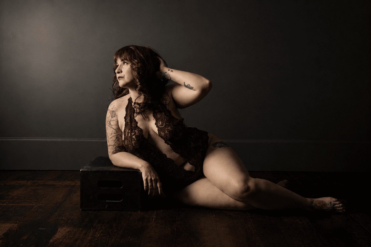 Woman in black lace lingerie reclining on a wooden floor against a dark backdrop during a fine art boudoir photography session in Dallas.