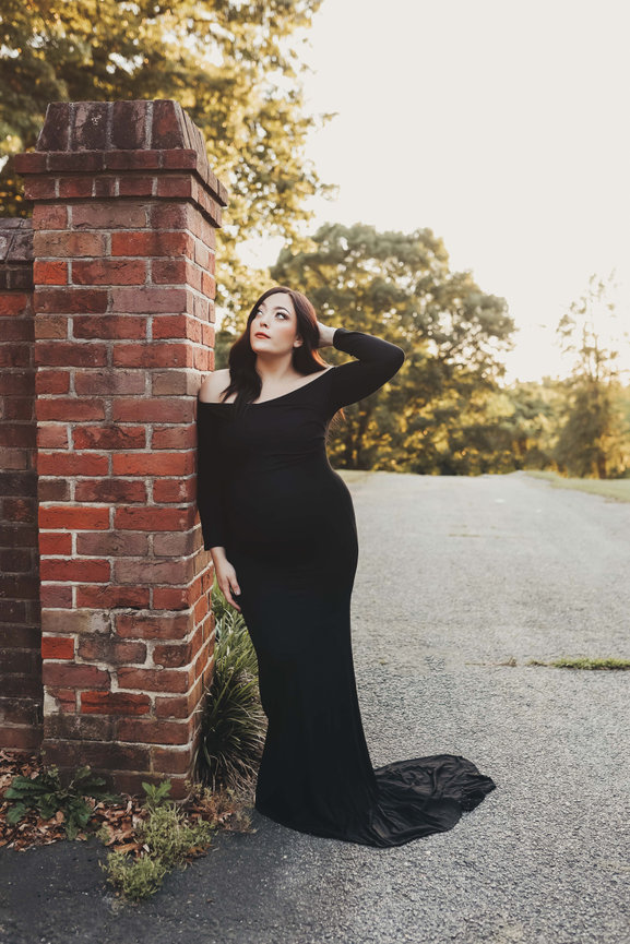 Woman in a black gown posing by a brick pillar on a tree-lined road at sunset