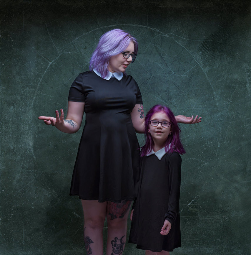 Adult and child with purple hair, glasses, and black dresses stand against a textured green background.