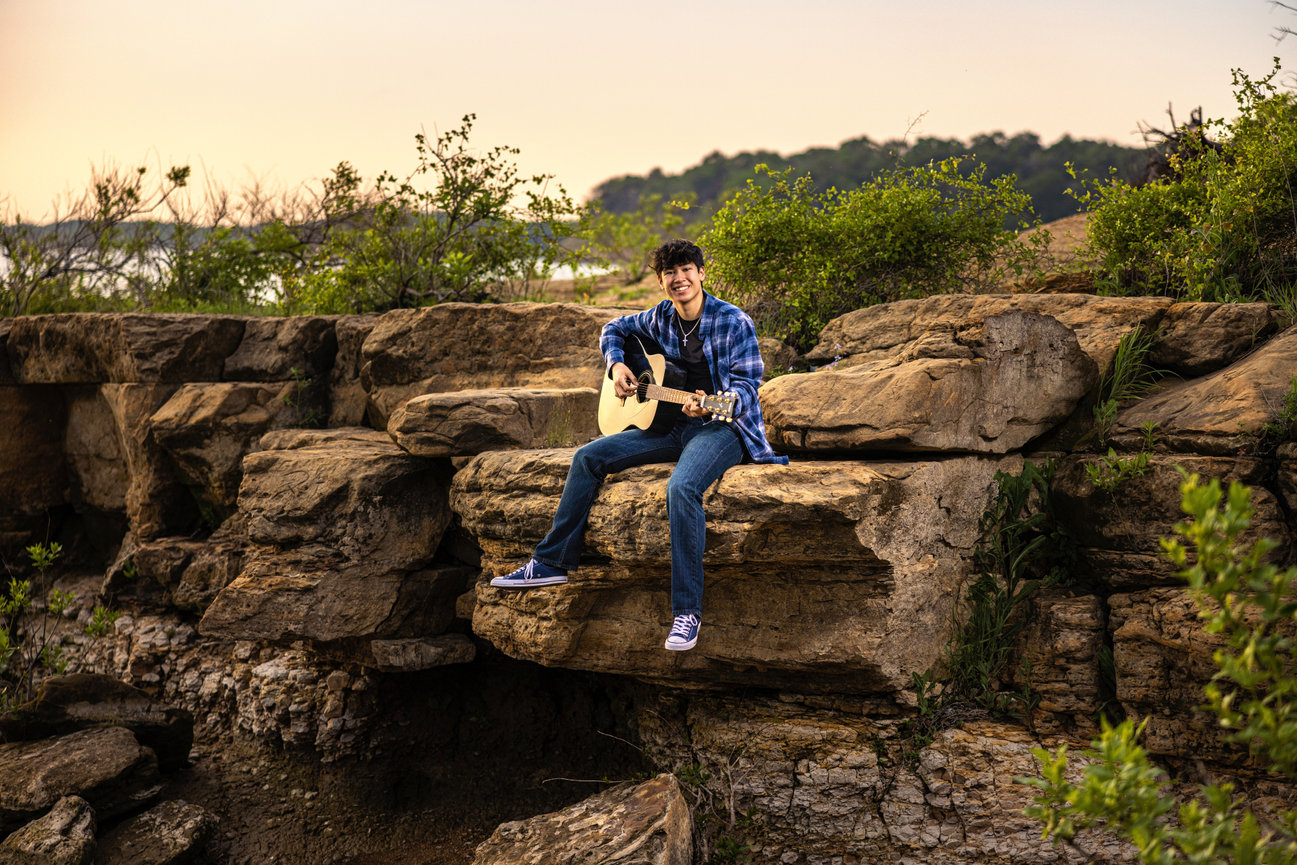 High school senior guy wearing jeans and a blue flannel shirt sitting on a rock playing his guitar as the sun sets.