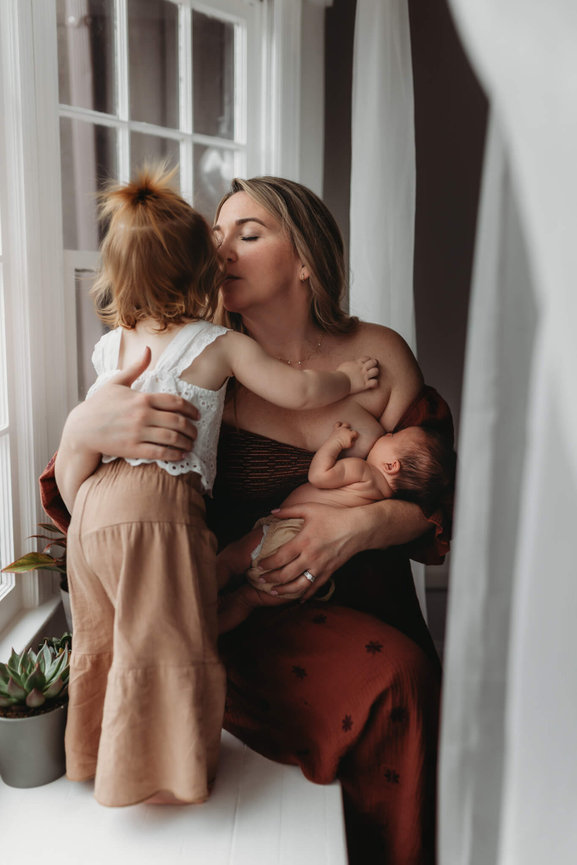 A mother sits in a window and kisses her daughter while nursing her newborn at a photography studio in Mendham, New Jersey.