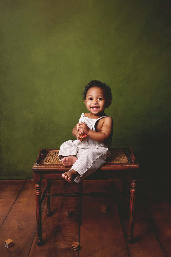 a laughing african-american boy holds blocks while sitting on a bench. The wall is pond green and the floor is wide plank wood.