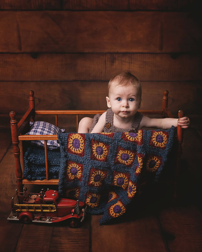 a six month old poses in a wood doll crib draped with a bright granny square blanket. There is toy firetruck in front. The floor and wall are wide plank wood.