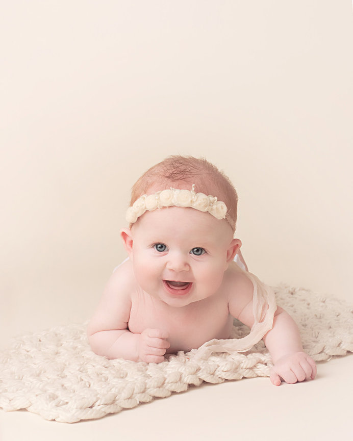 A grinning four month old baby pushes up on a cream knitted blanket on a beige backdrop