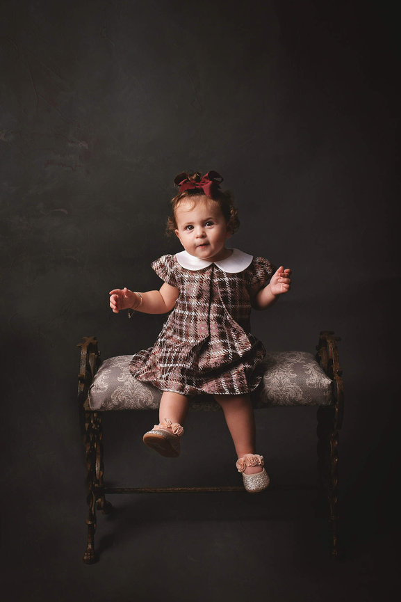 A one year old in a burgundy plaid dress swings her feet on an iron bench