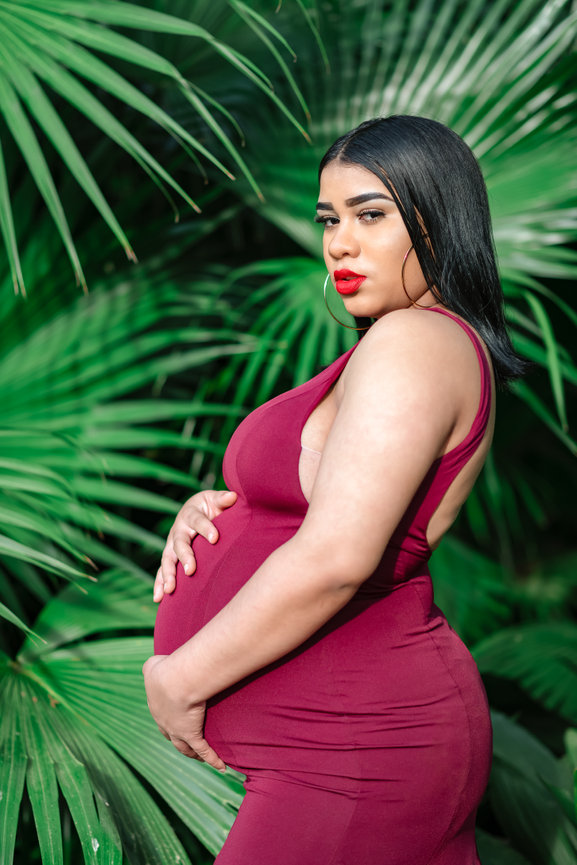 Pregnant woman in a red dress, standing in front of lush green palm leaves, gently holding her belly.