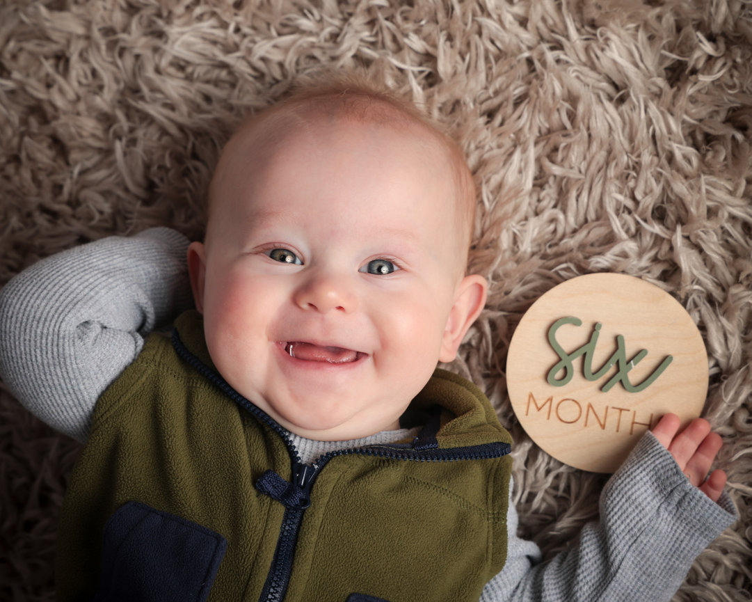 Smiling baby lying on a shaggy carpet holding a wooden sign reading Six Months.