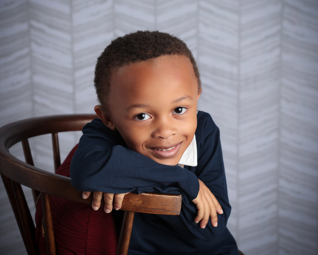 Smiling child leaning on a wooden chair, with a geometric-patterned background.