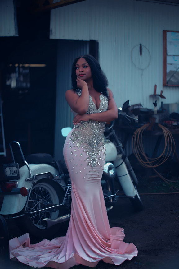 Woman in an elegant pink gown stands thoughtfully by a vintage motorcycle in an industrial setting.
