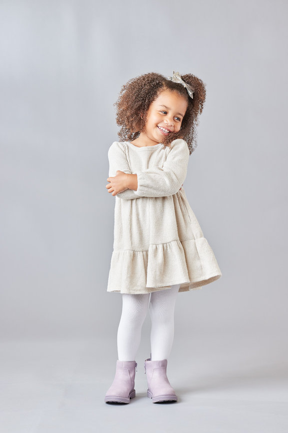 Full-body indoor portrait of a young child standing against a gray background, captured in soft natural-style light with a joyful and relaxed expression