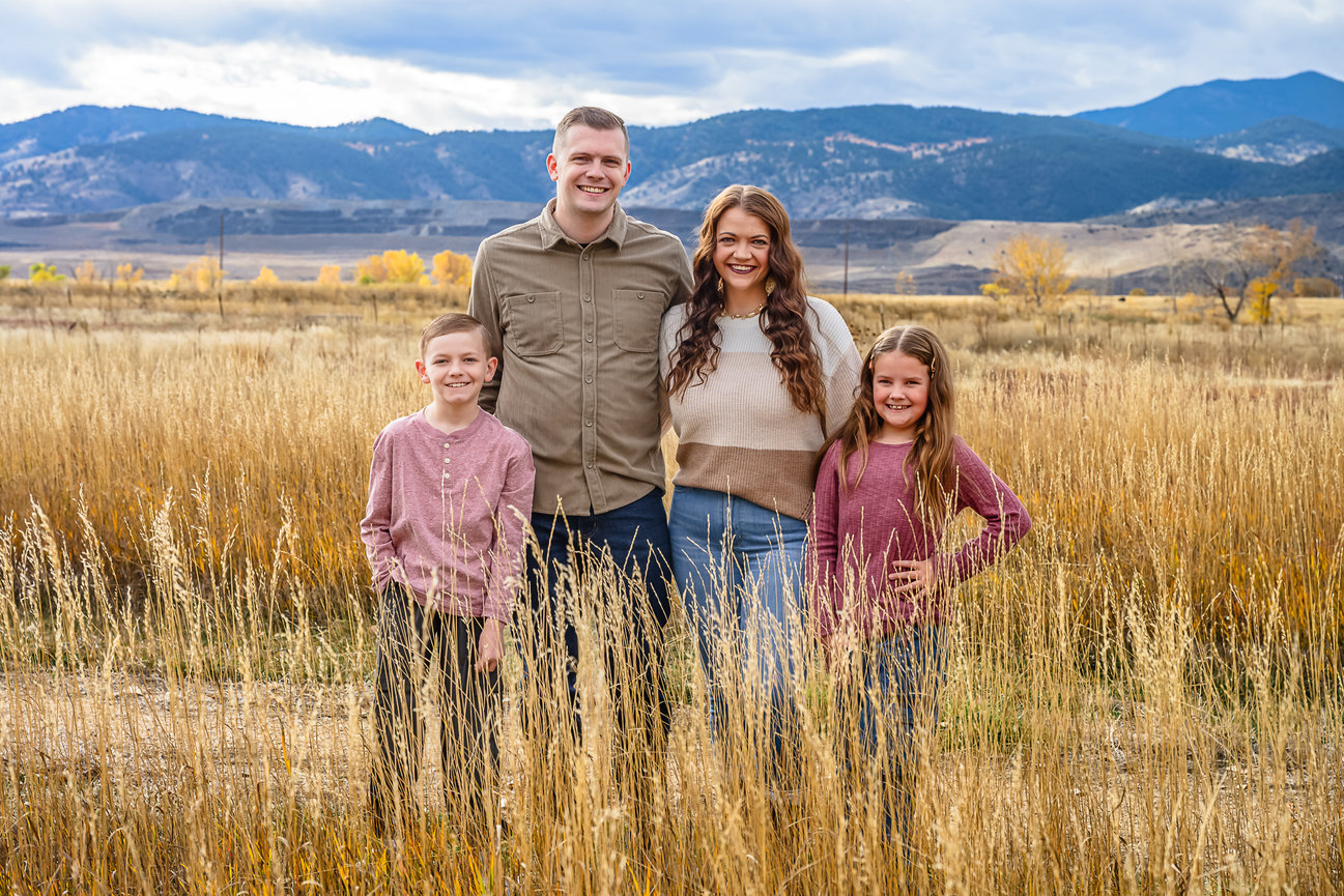 A gorgeous family of four stands in the tall grasses. The mountains are blue behind them.