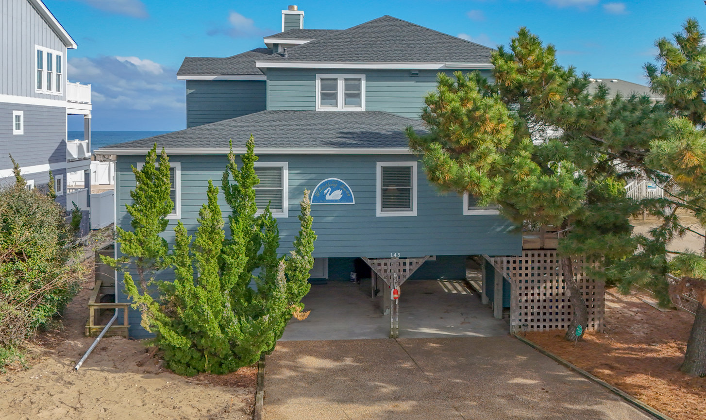 Two-story beach house with blue siding, elevated on stilts, with surrounding greenery and a clear sky backdrop.