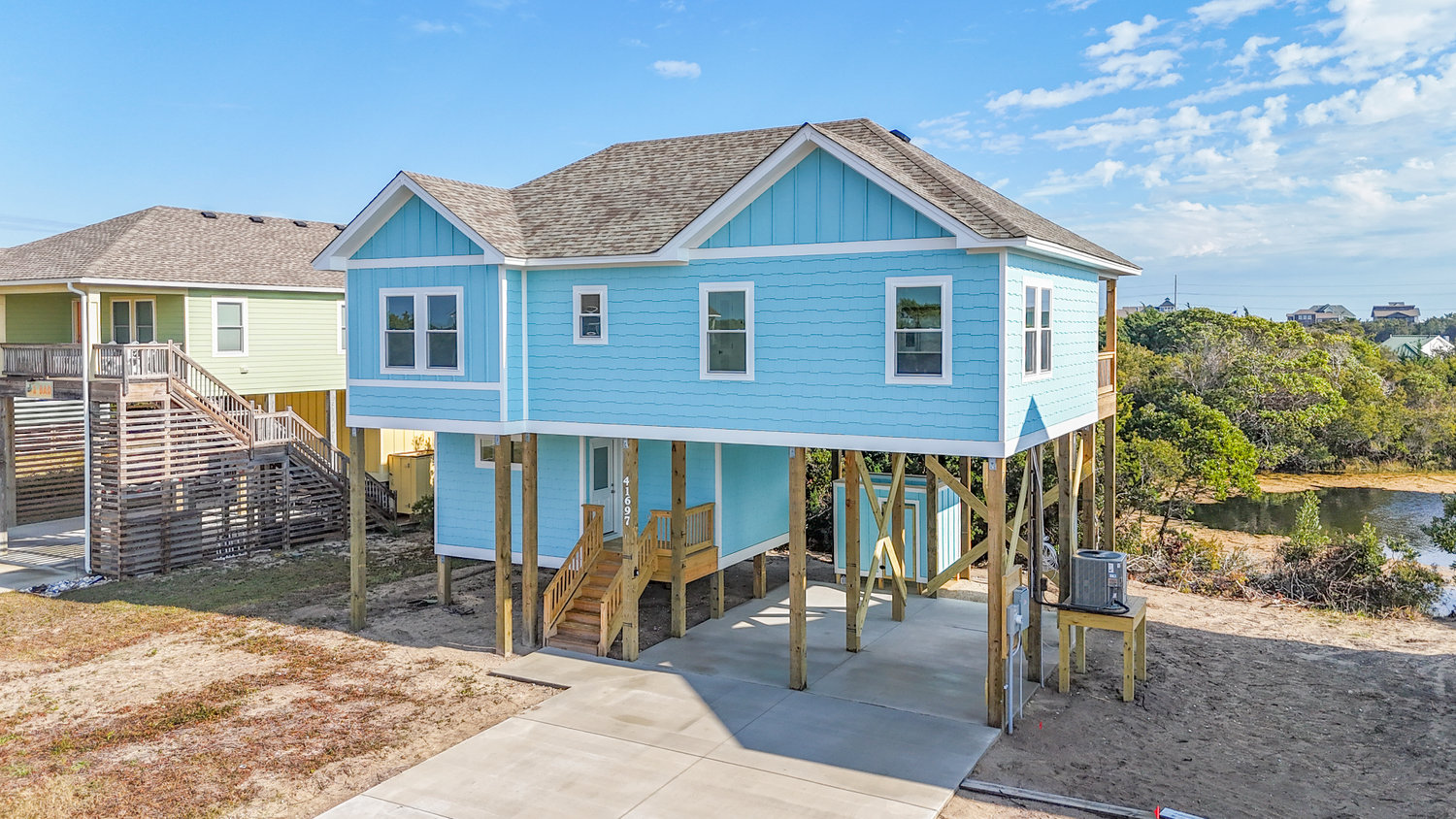 Elevated blue beach house on stilts with a driveway, surrounded by sand and trees, under a partly cloudy sky.
