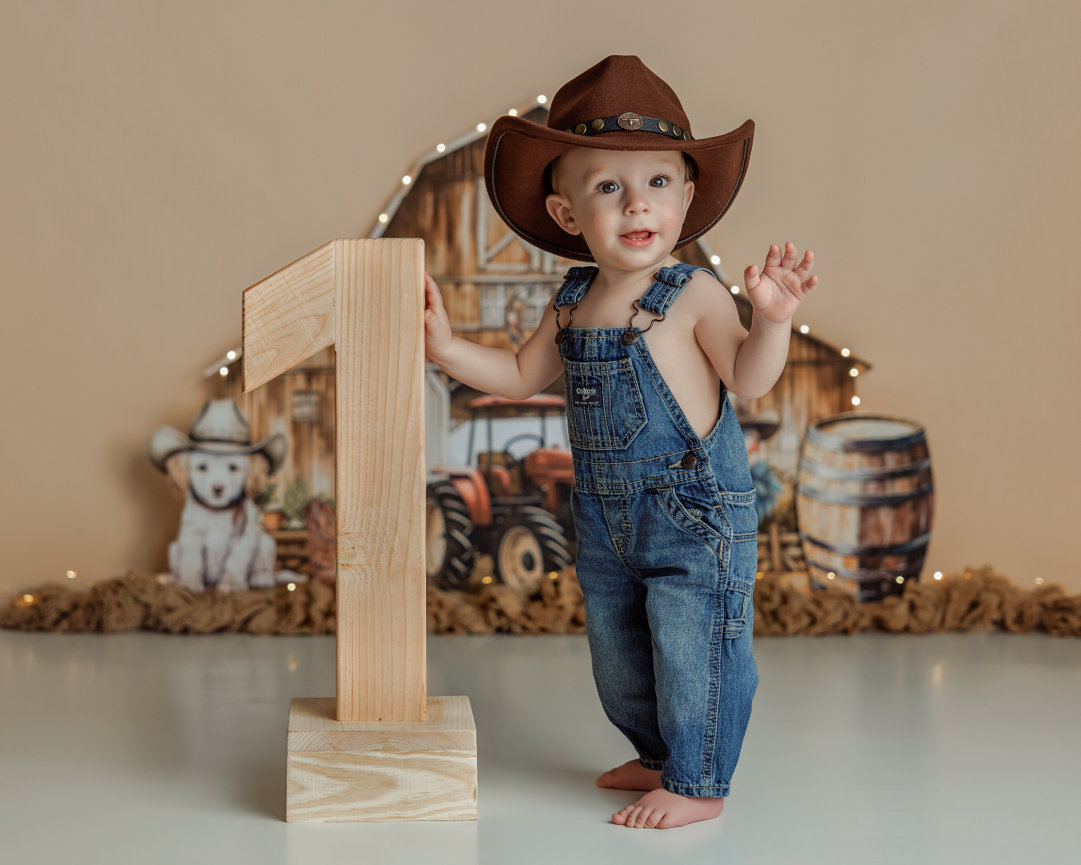 Toddler in overalls and a cowboy hat standing beside a barn-themed sign