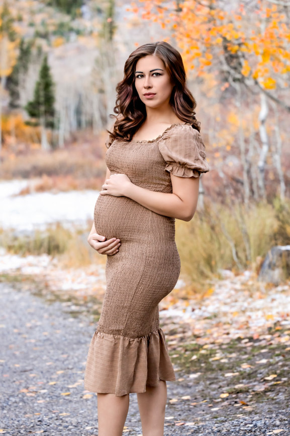 Pregnant woman in a brown dress standing on a path surrounded by autumn foliage.