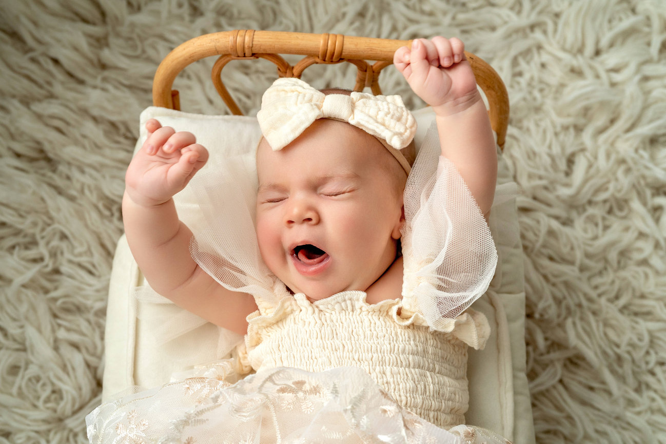 Sleeping baby yawning in a cream dress with a bow, lying in a wicker cradle on a fluffy rug