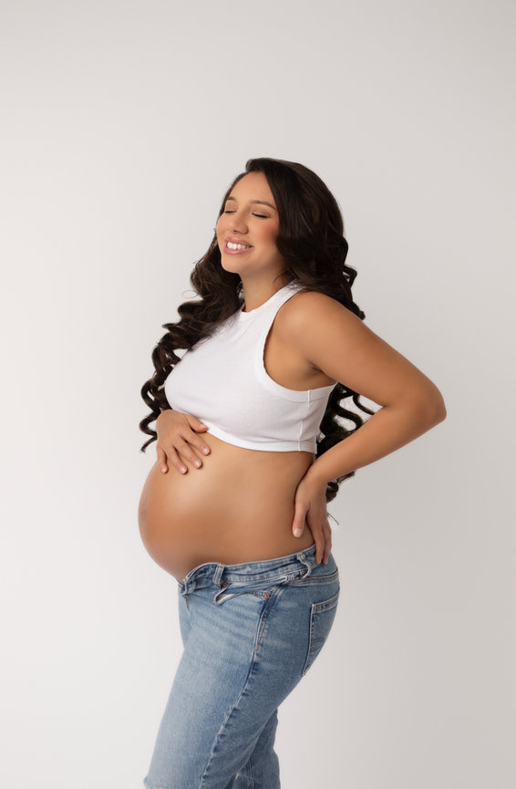 Pregnant woman in a white tank top and jeans, holding her belly, smiling.